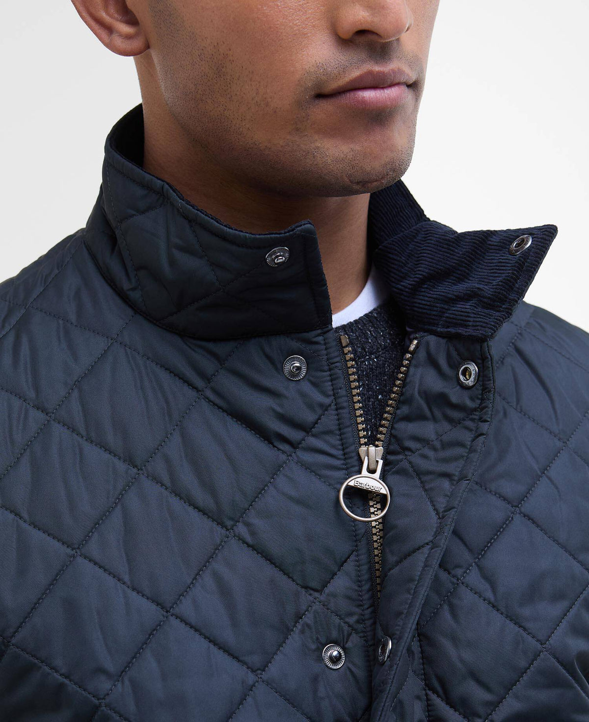 Man wearing a quilted navy jacket with the zip open a small way. Cord fabric lining inside the collar, button popers and a Barbour branded zip pull