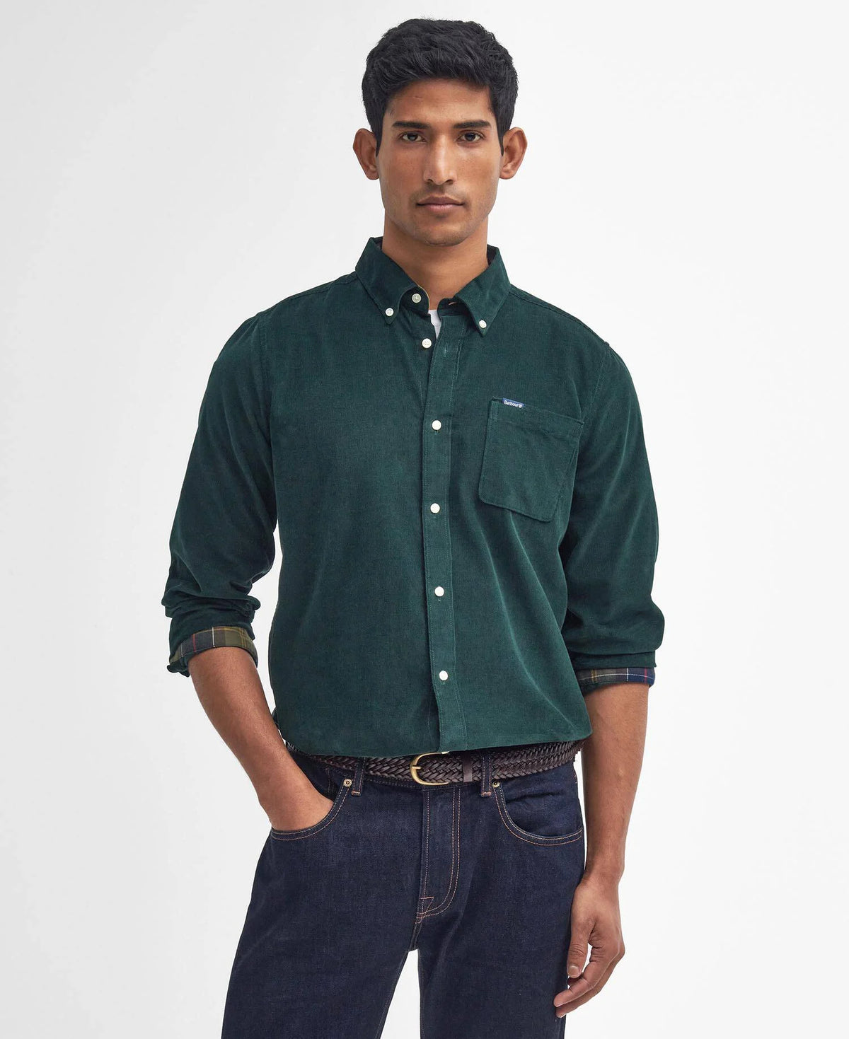 A man wearing a dark green corduroy button-down shirt with a chest pocket, paired with dark blue jeans, standing against a light gray studio background.