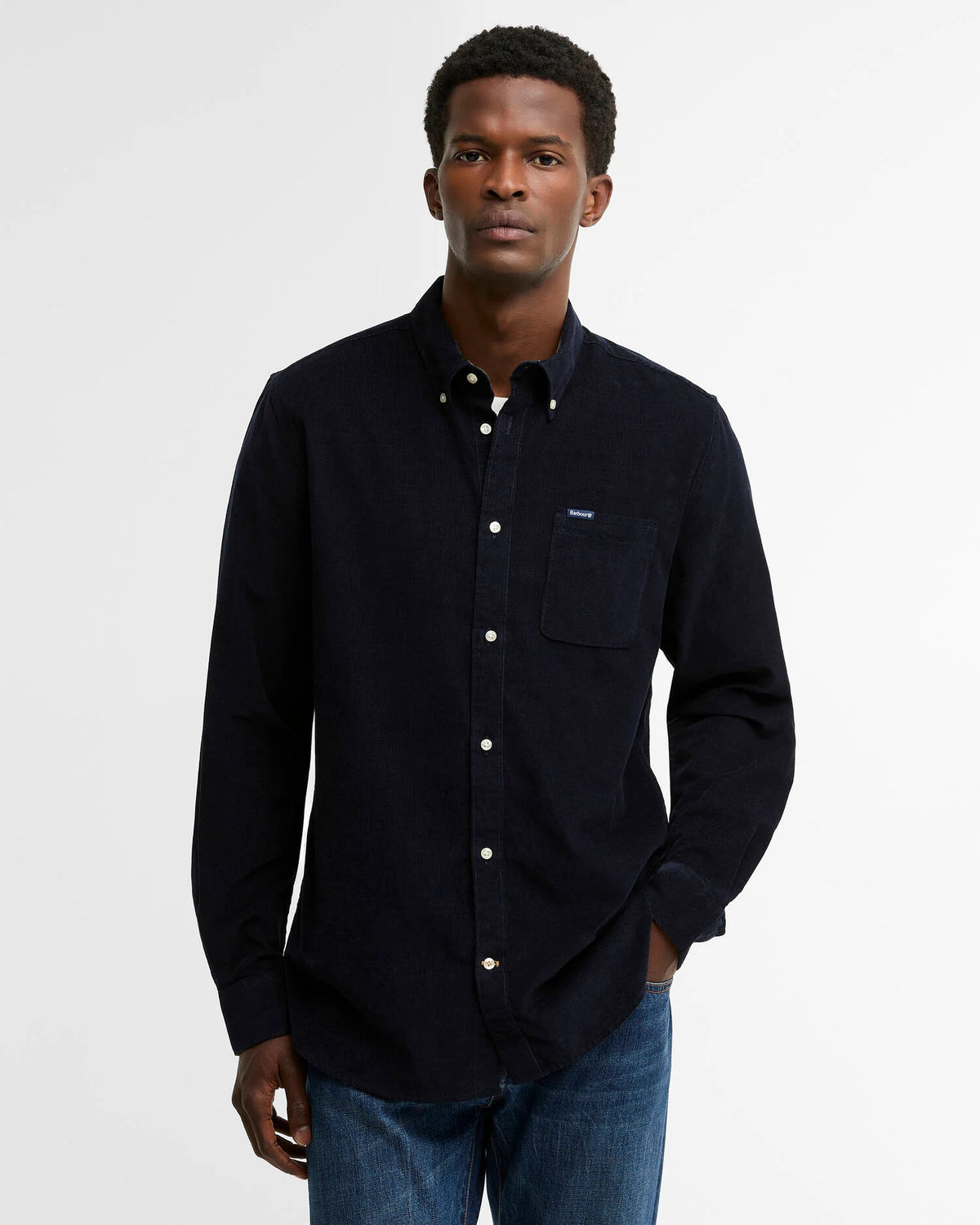 A man wearing a dark navy long-sleeve button-down shirt with a chest pocket, paired with blue jeans, photographed against a plain light gray studio background.