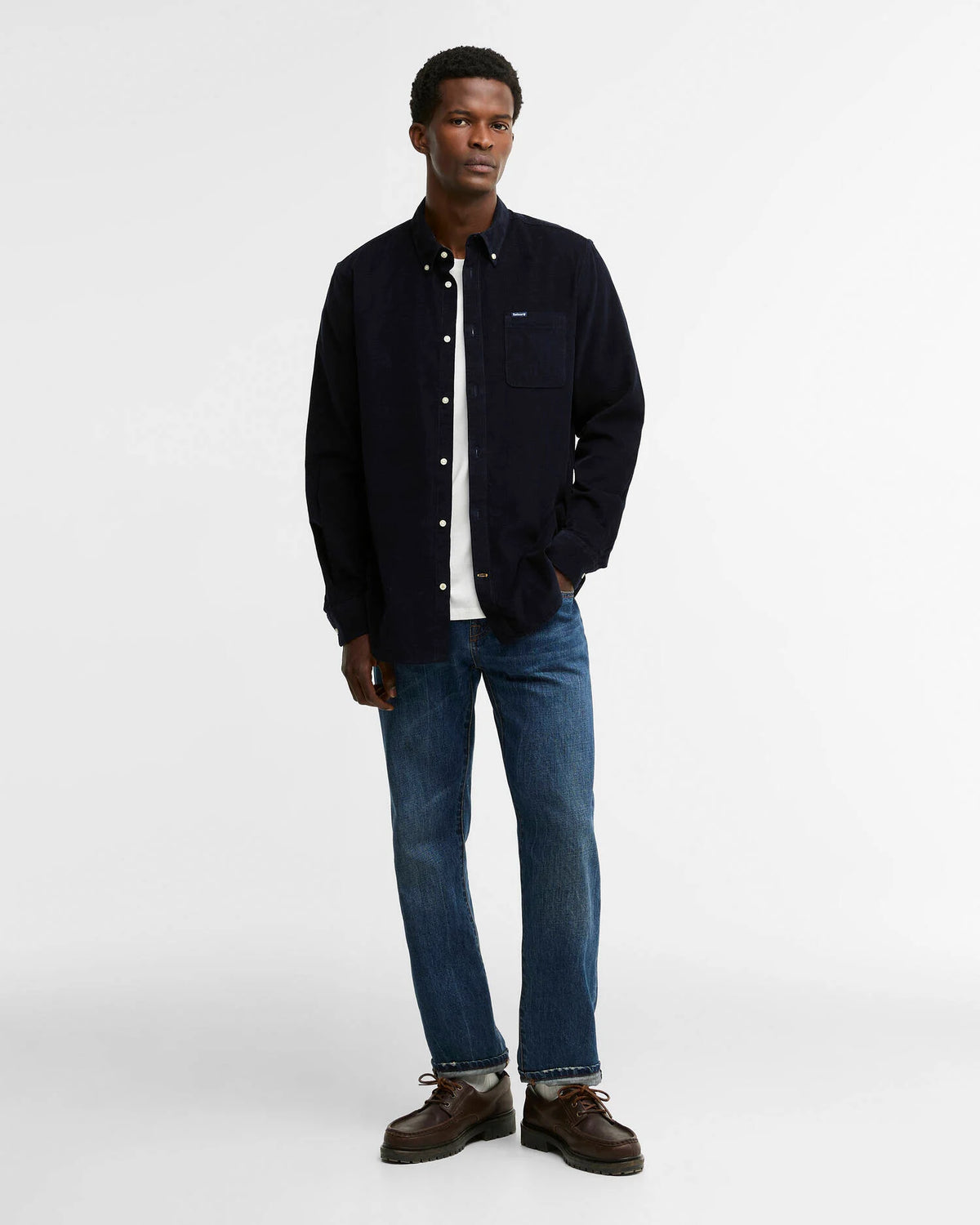 A man modeling a dark navy blue button-up cord shirt over a white t-shirt, paired with blue jeans and brown leather shoes, against a plain light gray background.