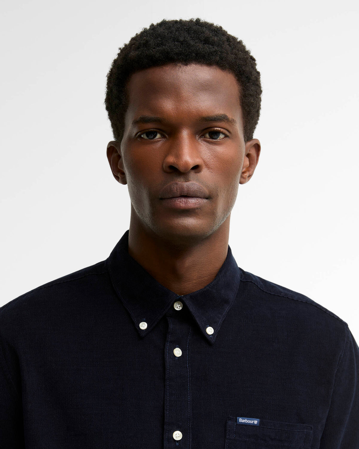 A man wearing a dark navy blue button-down cord shirt by Barbour, photographed against a clean light gray background.