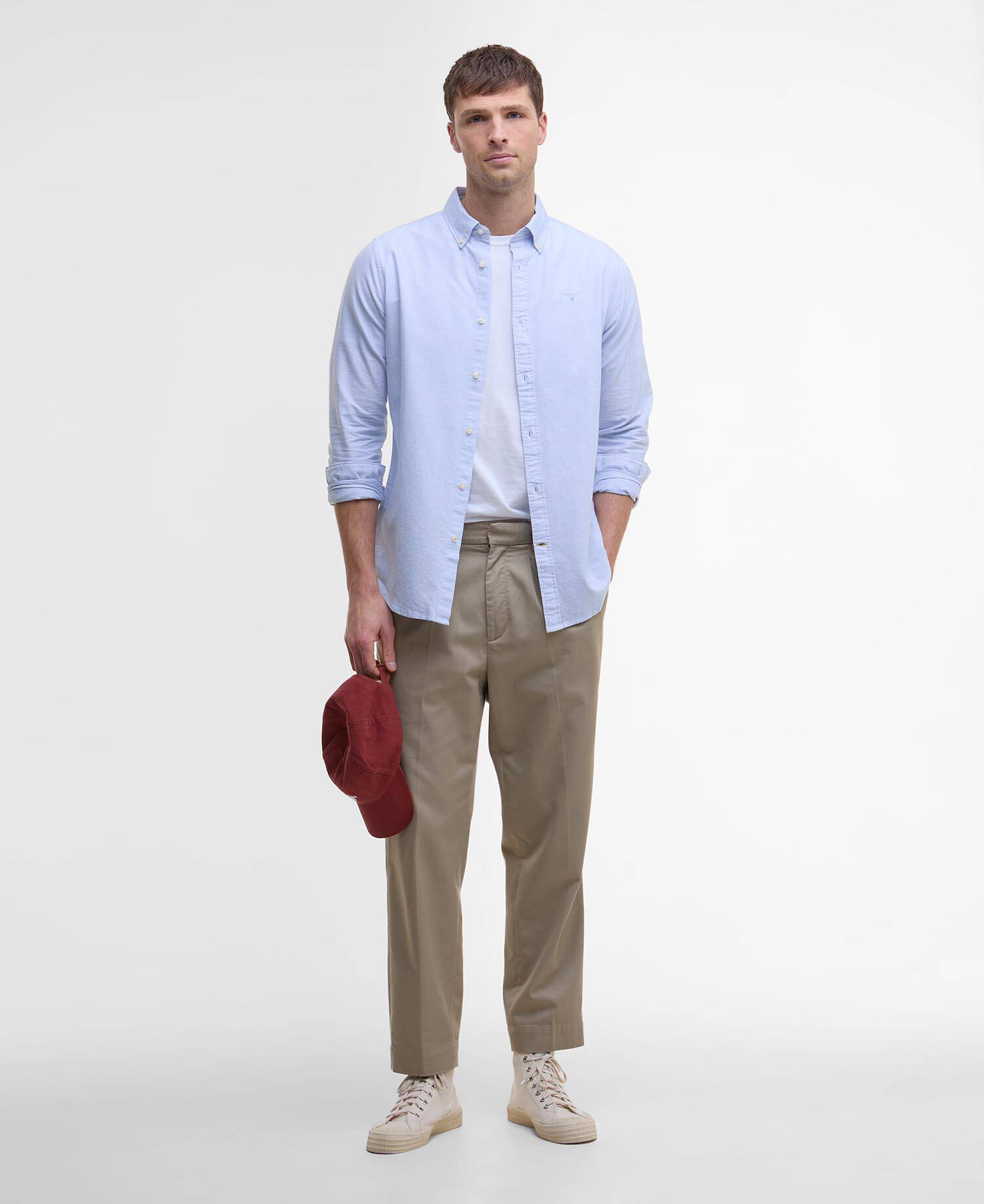 Man wearing a light blue shirt, beige pants, and beige shoes, holding a red hat on a white background