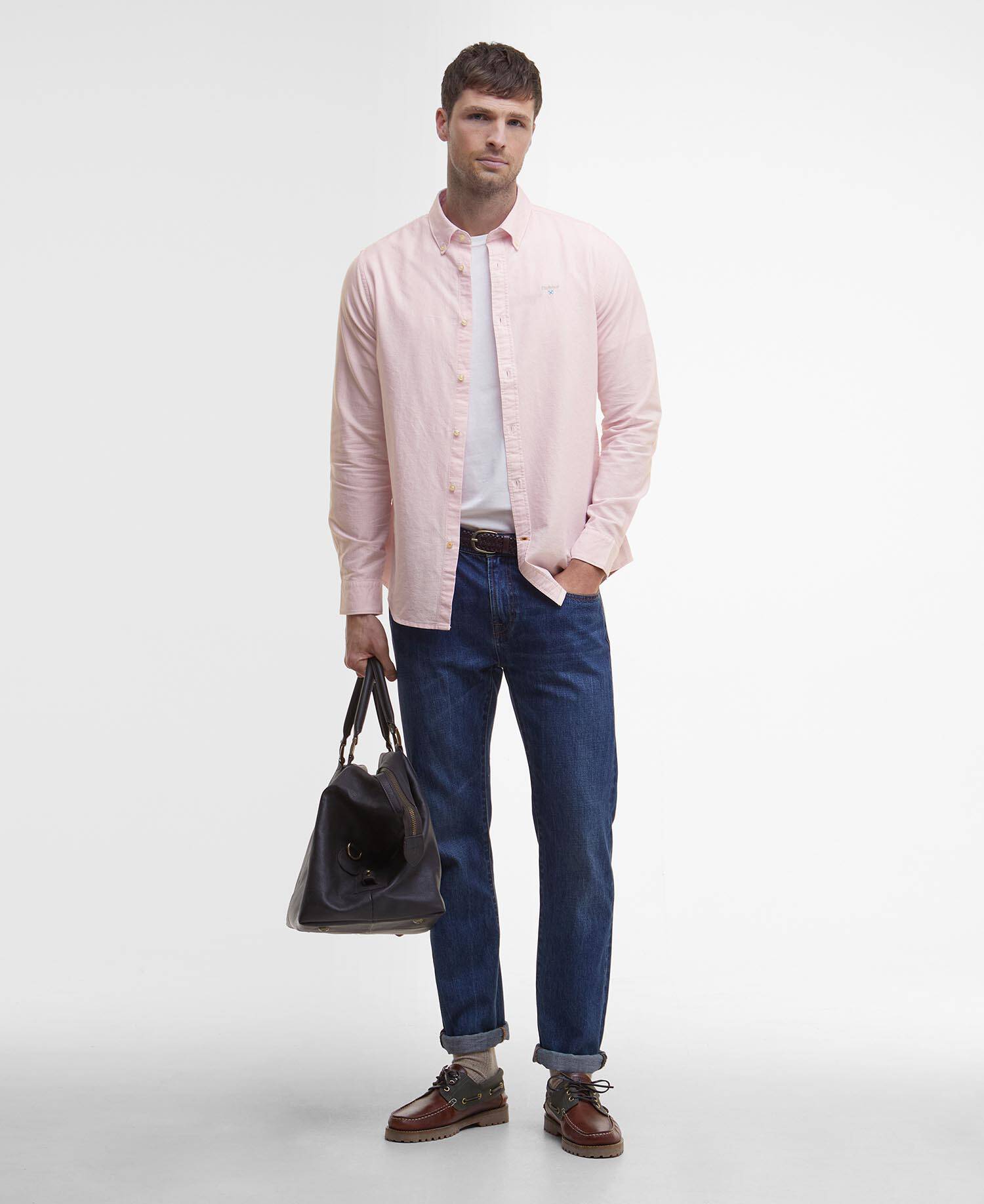 Man wearing a light pink Barbour shirt, blue jeans, white T-shirt and brown shoes and holding a leather bag