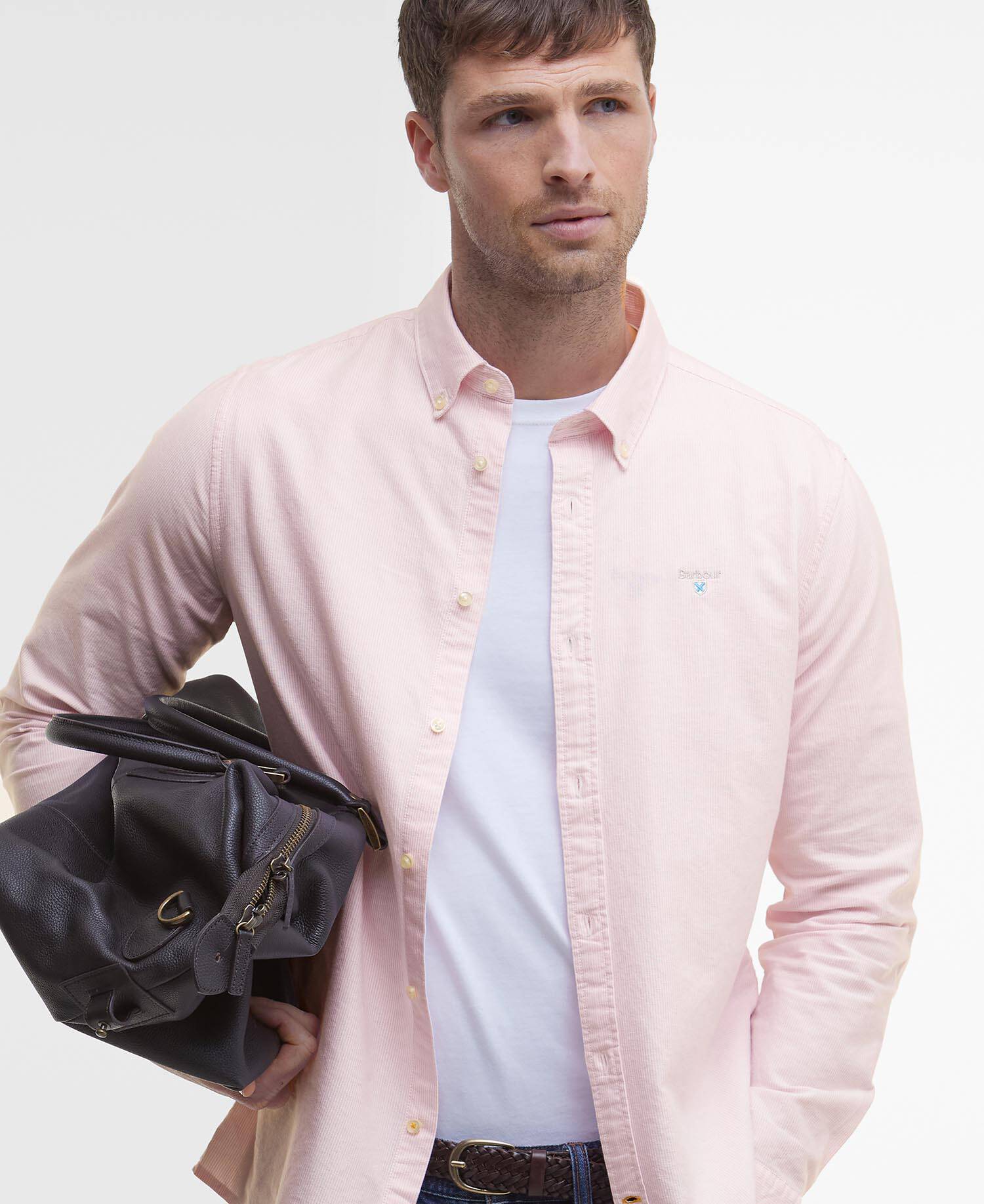 Man wearing a light pink shirt over a white t-shirt, holding a black bag against a plain background