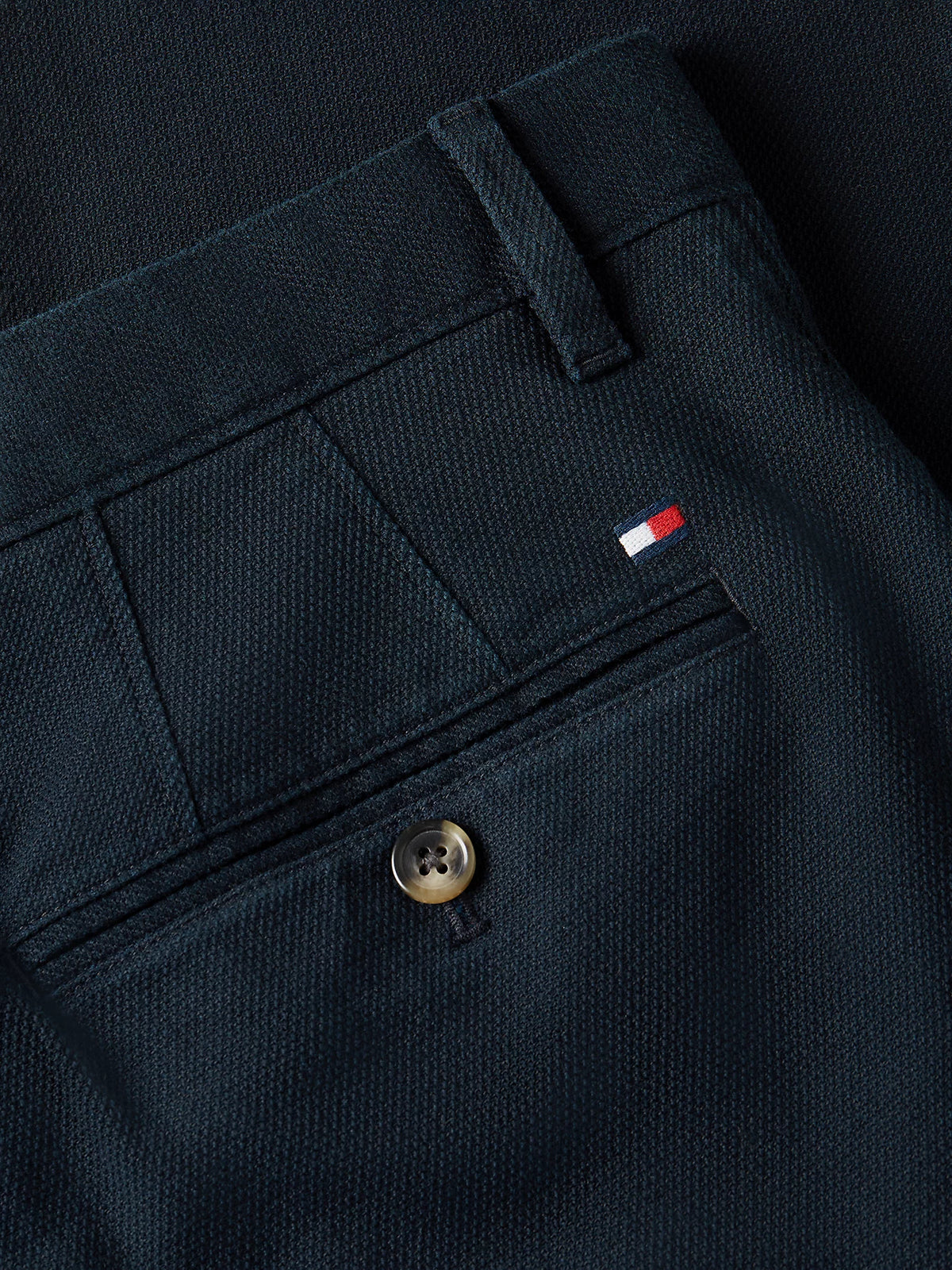 Close-up of dark navy trousers showing the back pocket with a button closure and a small Tommy Hilfiger flag logo tab.