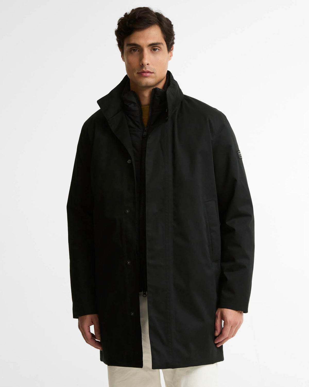A man wearing a black mid-length parka jacket with a high collar, shown from the waist up against a plain light gray studio background.