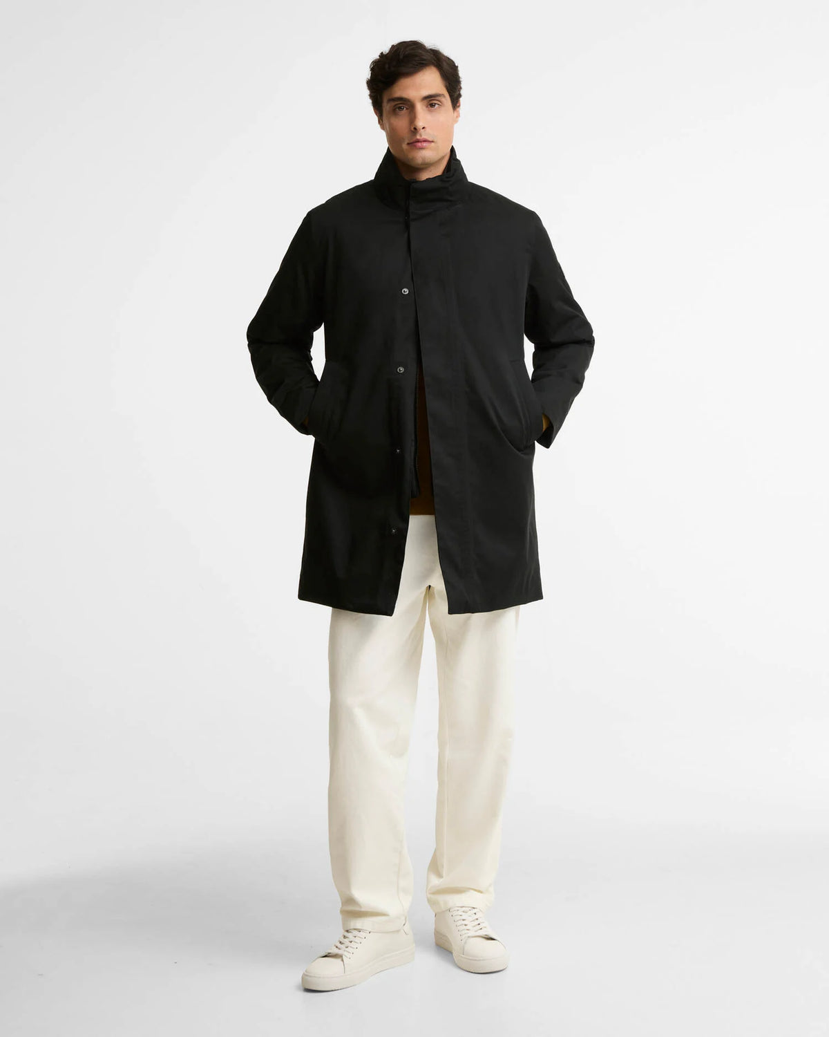 A man modeling a black mid-length coat with a high collar, worn over a brown shirt and cream trousers, standing against a light gray studio background.