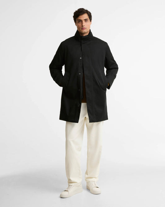 A man modeling a black mid-length coat with a high collar, worn over a brown shirt and cream trousers, standing against a light gray studio background.
