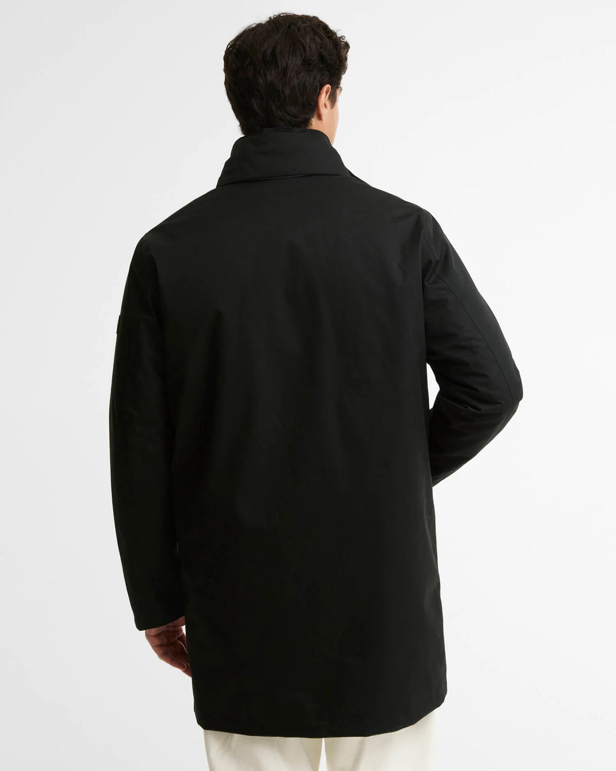 A man viewed from behind wearing a black mid-length jacket with a high collar and cream pants, set against a plain white studio background.