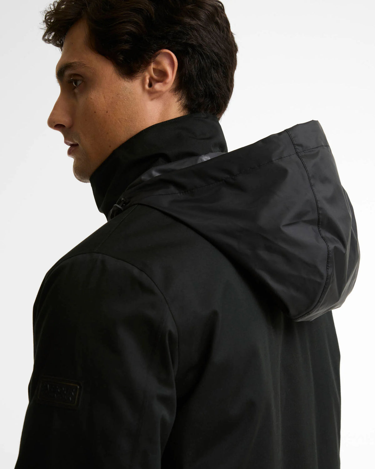 A man shown in profile wearing a black hooded jacket against a plain light gray studio background.