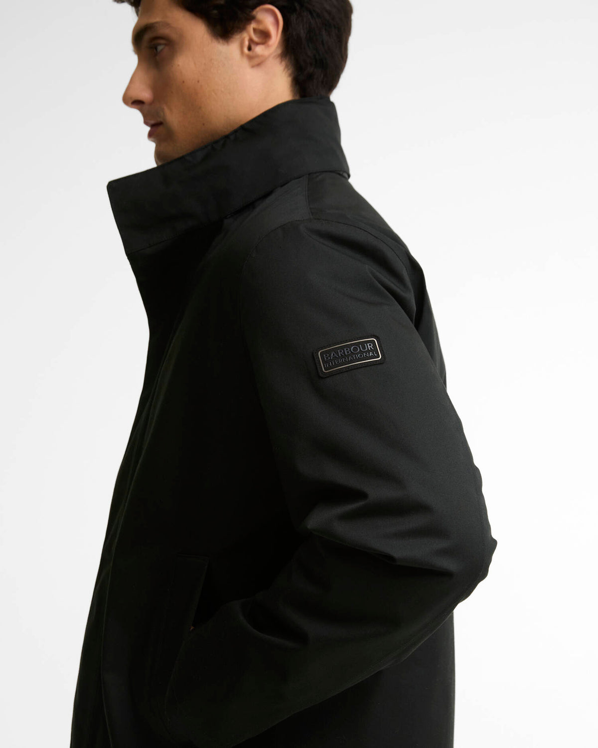A man in profile wearing a black Barbour International jacket with a high collar and a visible brand patch on the upper sleeve, set against a plain light gray background.