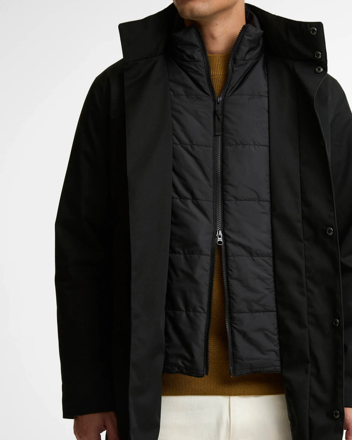 A close-up of a person wearing a black outerwear jacket with a high collar over a black quilted zip-front lining, layered over a mustard-brown sweater and white pants, against a plain light background.