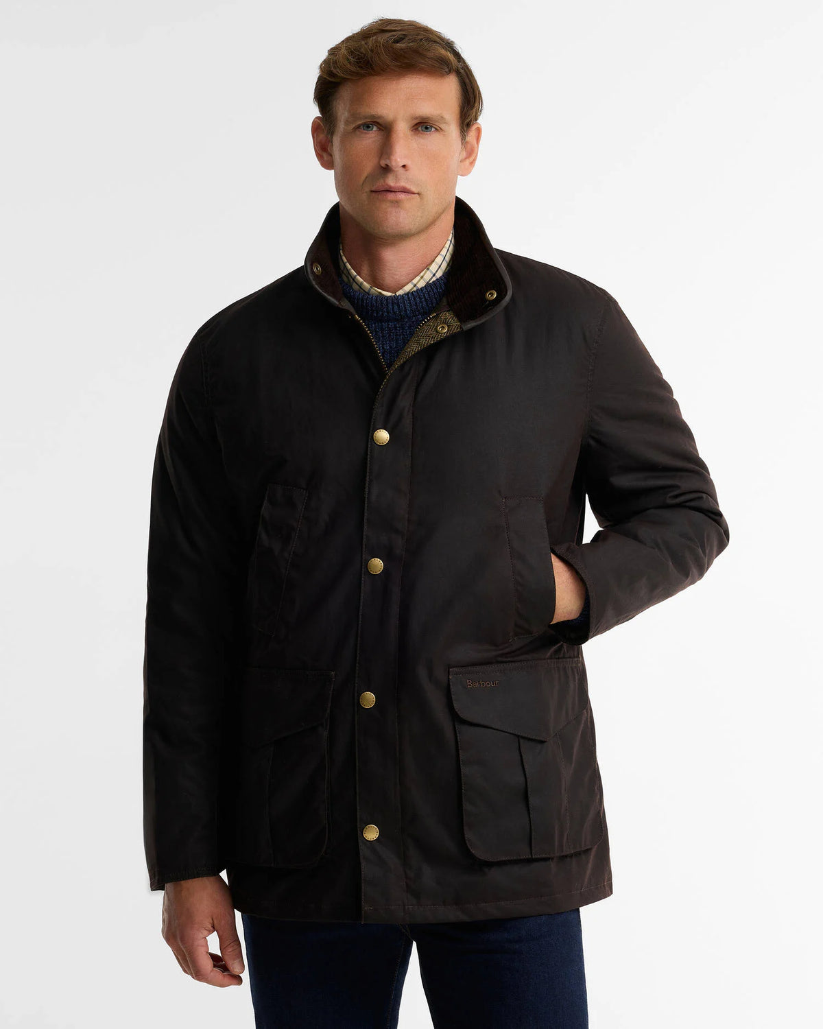 A man wearing a dark brown waxed cotton field jacket with gold-tone snap buttons and a brown corduroy collar, layered over a blue knit sweater and dark trousers, photographed against a light gray studio background.