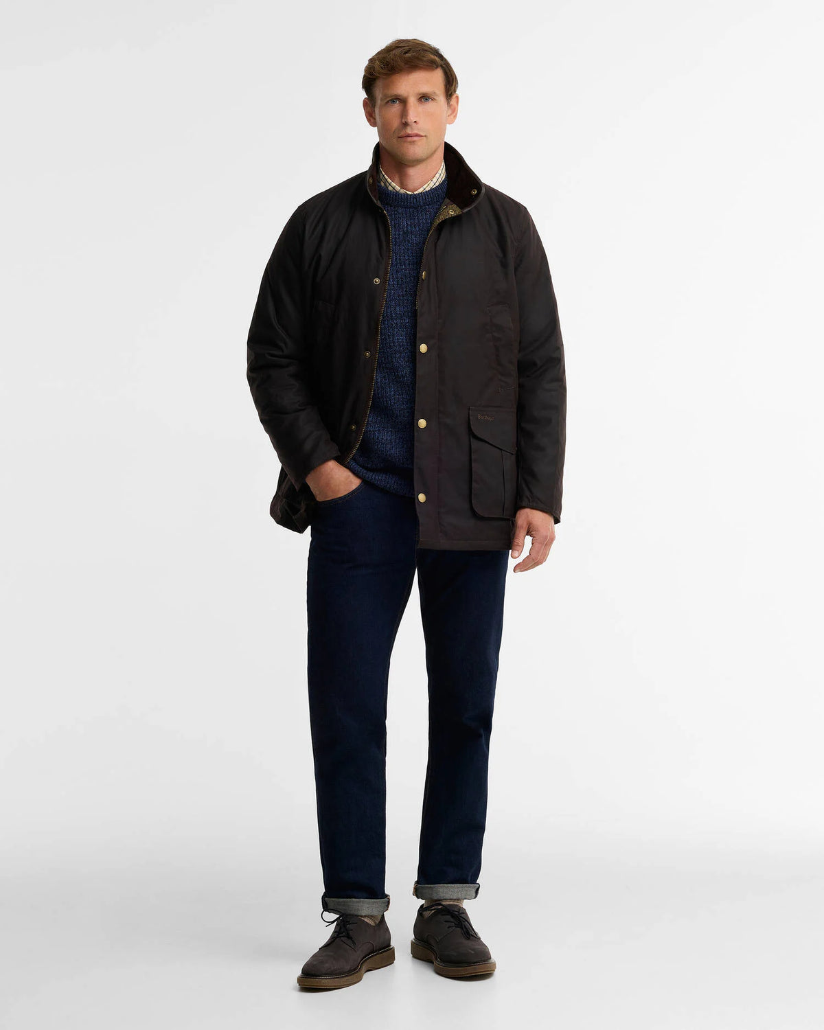 A man modeling a dark waxed cotton jacket with brass buttons, worn over a navy knit sweater, dark blue jeans, and brown shoes, photographed against a clean white studio background.