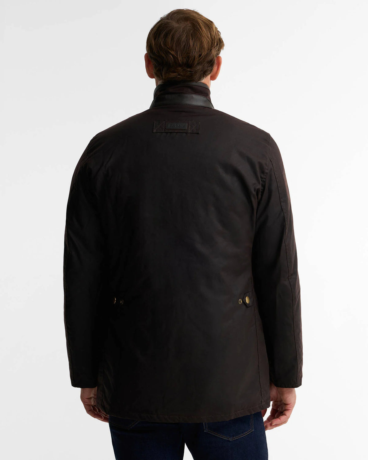 A man viewed from the back wearing a dark brown waxed jacket and blue jeans, standing against a plain white studio background.