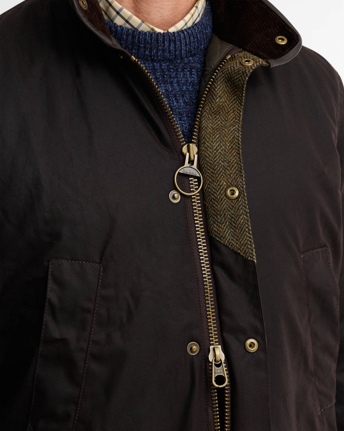 Close-up of a dark brown waxed cotton jacket with a brass zipper, showing the interior herringbone lining and a blue knit sweater underneath.