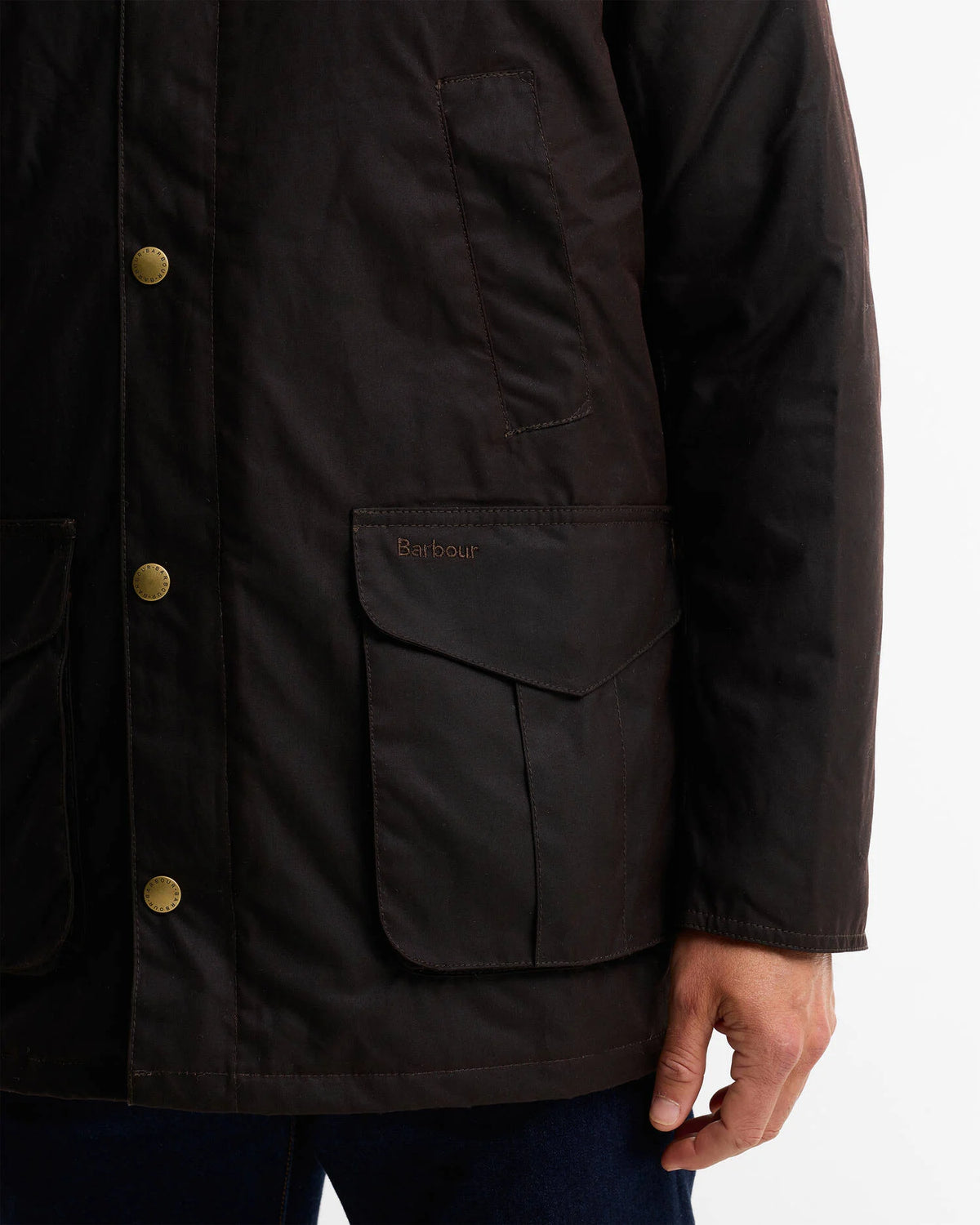 Close-up of a person wearing a dark brown waxed jacket with brass buttons and a front flap pocket, with the Barbour logo embroidered on the pocket.