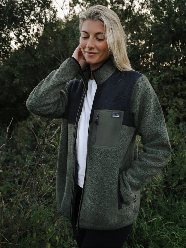 A woman wearing an olive green and black fleece jacket with a white t-shirt underneath, standing outdoors in a wooded setting.