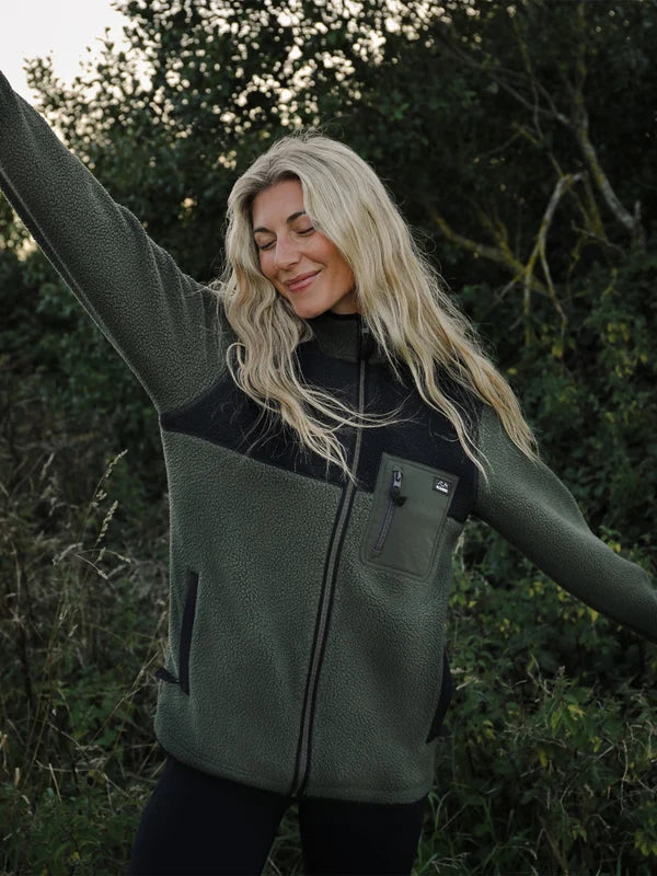 A woman outdoors wearing an olive green fleece jacket with black panels and a front zipper, posing with her arms outstretched against a backdrop of trees and foliage.