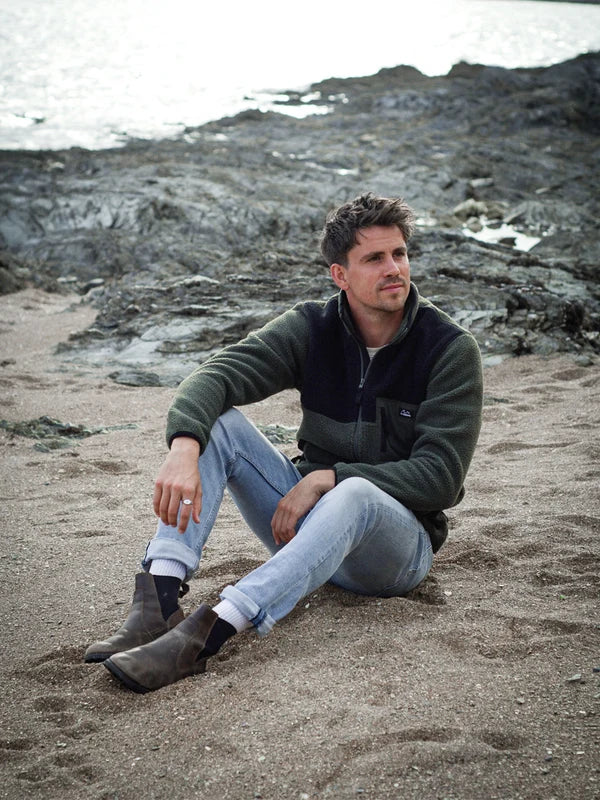 A man sits on a sandy beach with rocky shoreline and water in the background, wearing a dark green and black full zip fleece jacket, light blue jeans, and dark brown ankle boots.