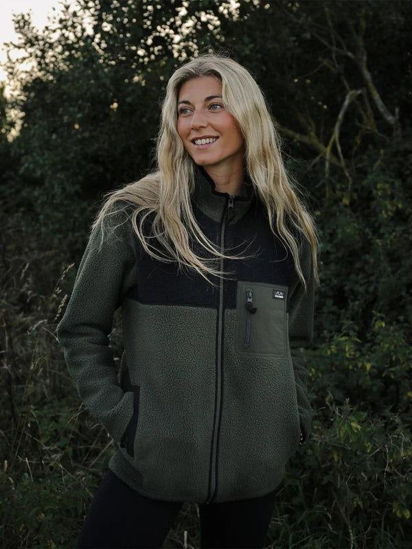 A woman with long blonde hair stands outdoors in a wooded setting wearing a color-block fleece jacket. The jacket has a dark olive green lower body and sleeves, with a black yoke and collar, and a front zipper.