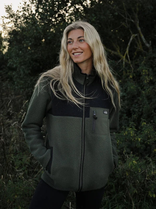 A woman with long blonde hair stands outdoors in a wooded setting wearing a color-block fleece jacket. The jacket has a dark olive green lower body and sleeves, with a black yoke and collar, and a front zipper.
