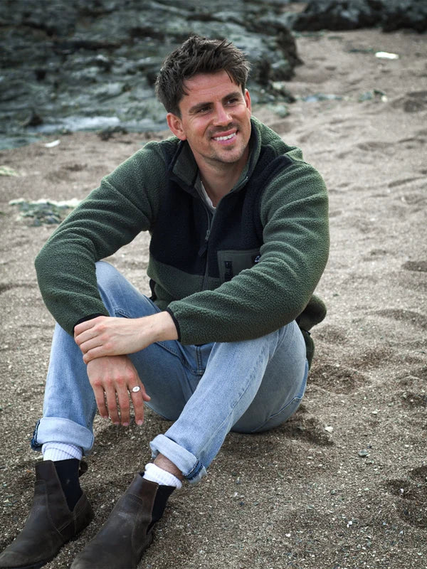 A man sitting on a sandy beach wearing a dark green fleece zip-up jacket over a black shirt, light blue jeans, and dark brown boots.