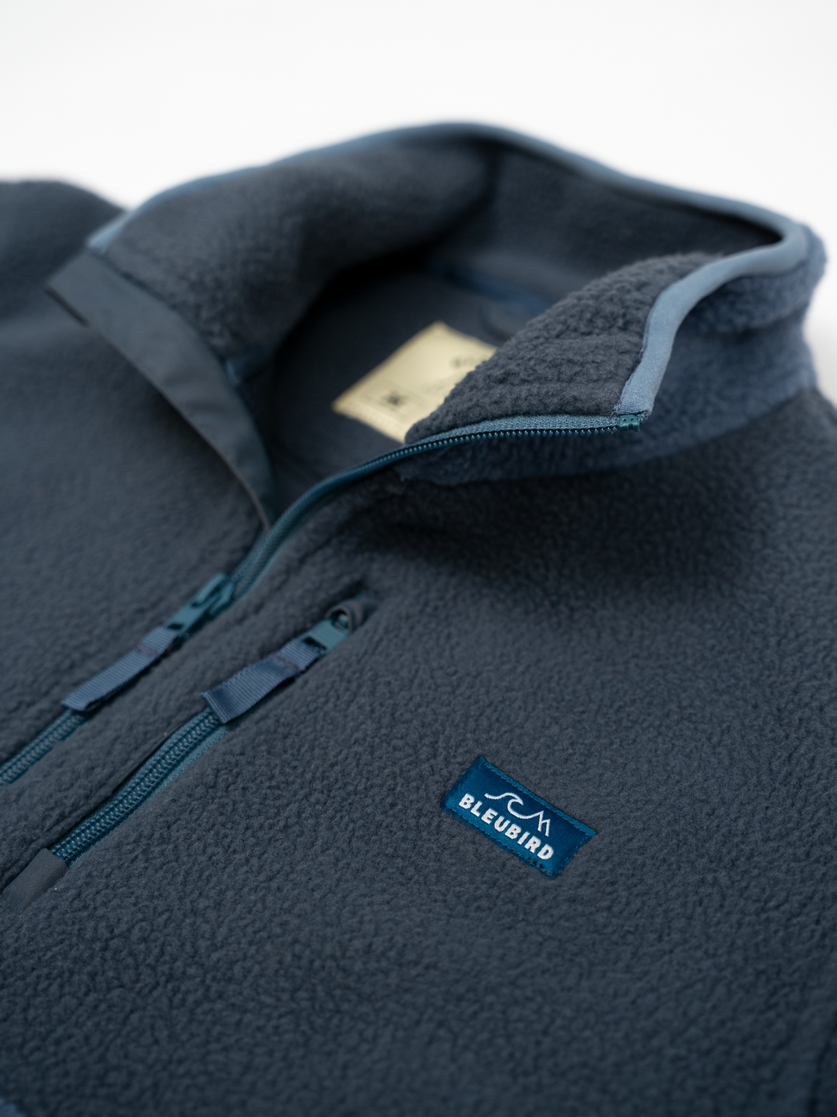 Close-up of a fleece pullover jacket in dark navy blue, showing the collar, zipper, and a small blue Bleubird logo patch on the chest.