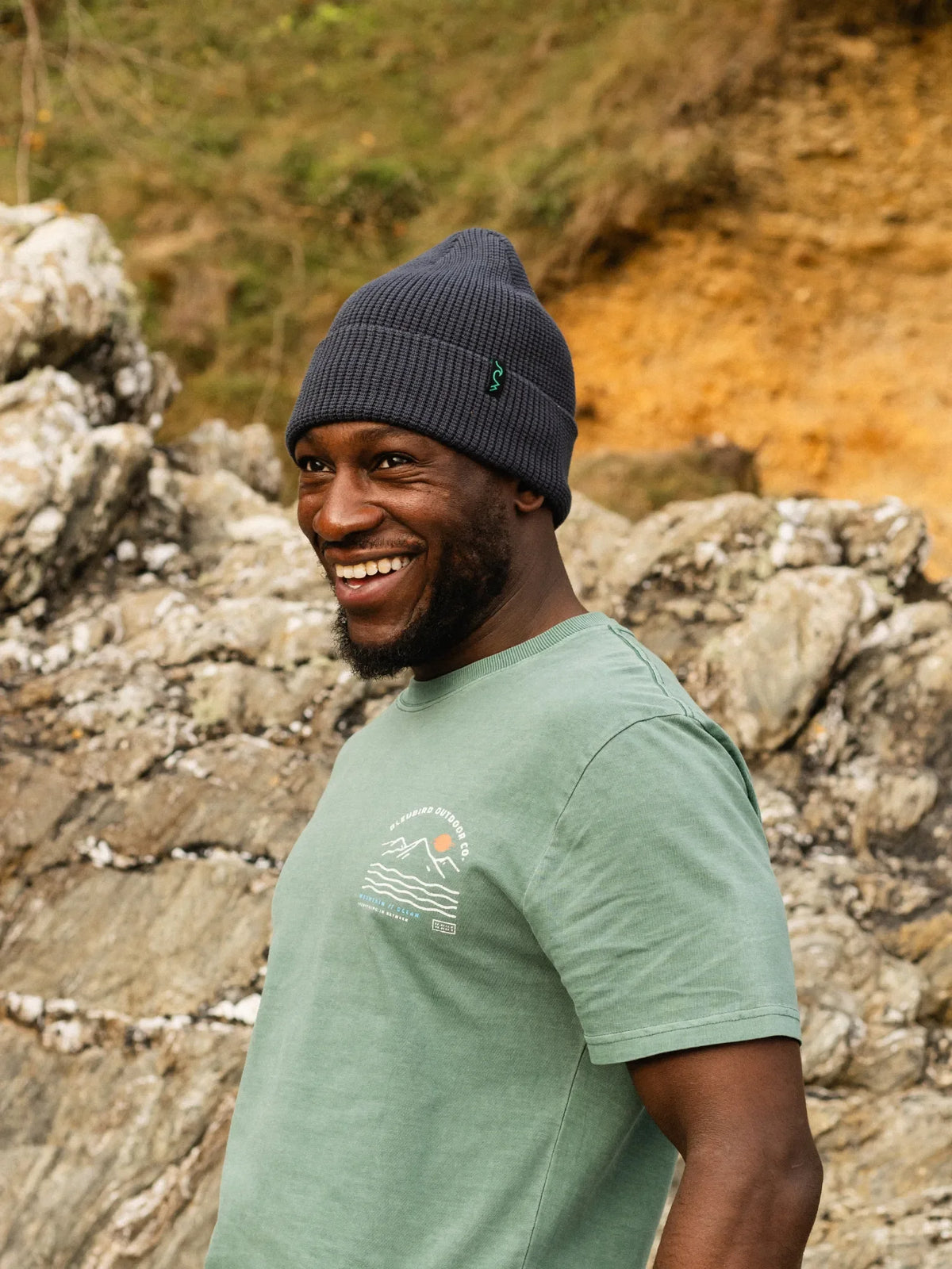 A man smiling outdoors in front of rocky terrain, wearing a dark gray ribbed beanie and a sage green t-shirt with a small mountain graphic on the chest.