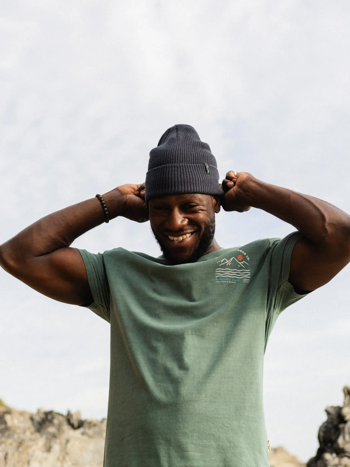 A smiling man wearing a dark gray knit beanie and a sage green t-shirt with a small mountain logo on the chest, posing outdoors against a cloudy sky.