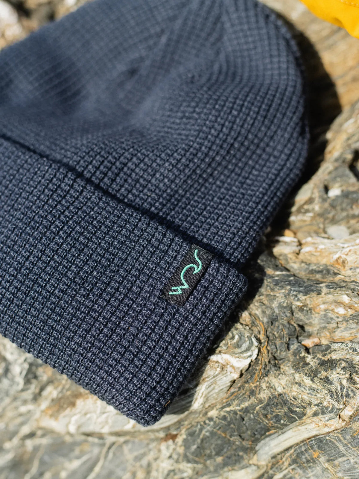 A close-up of a dark navy blue waffle-knit beanie resting on a textured gray stone surface. A small mint green embroidered stylized logo is visible on the cuff.