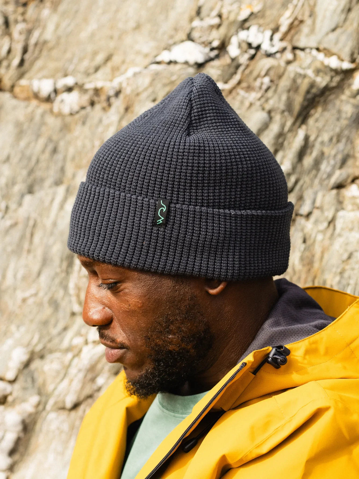 A man wearing a dark gray ribbed knit beanie with a small green embroidered logo, paired with a bright yellow jacket, set against a textured stone wall background.