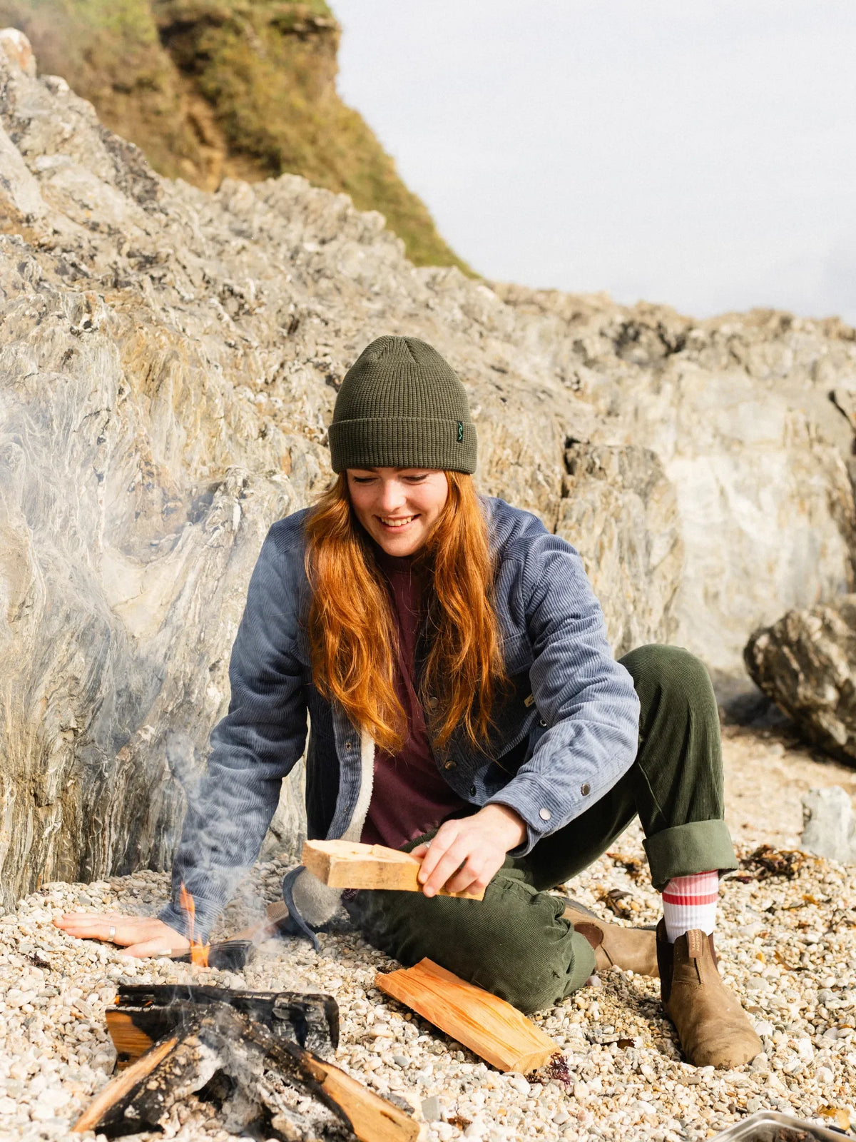 A woman with long red hair sits on a rocky beach, wearing an olive green beanie, a blue-gray fleece jacket, and dark green pants while lighting wood near a small fire.