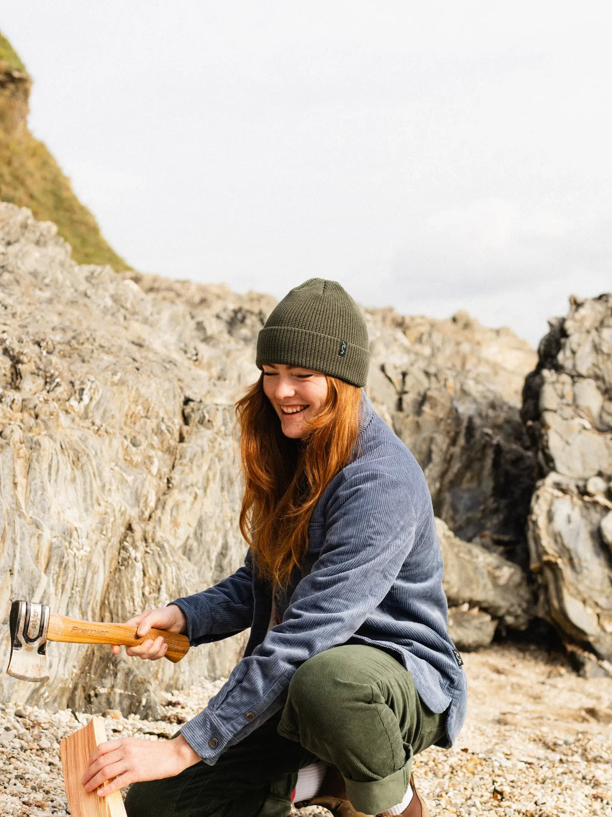 A woman crouching outdoors among large rocks, wearing an olive green beanie, a blue-gray fleece jacket, and olive pants, while holding a small axe and wood piece.