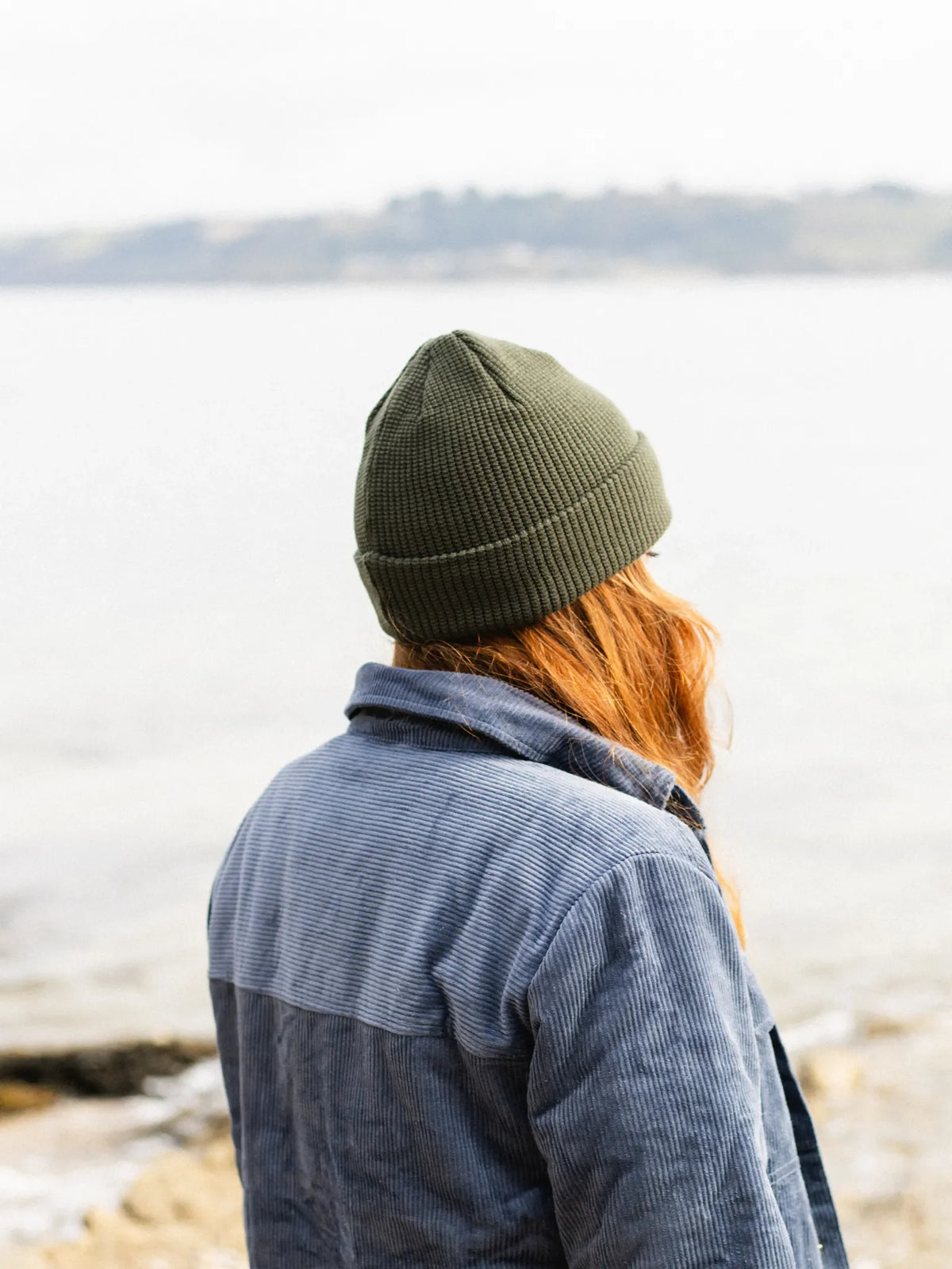 A person seen from behind wearing an olive green ribbed knit beanie and a blue corduroy jacket, standing outdoors near a body of water with a blurred shoreline in the background.