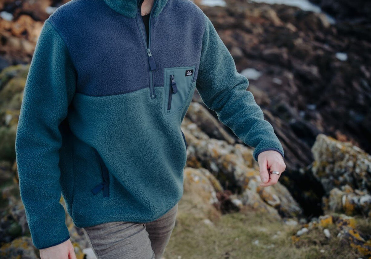 A person wearing a color-block fleece pullover in teal and navy, walking outdoors on a rocky, grassy path.