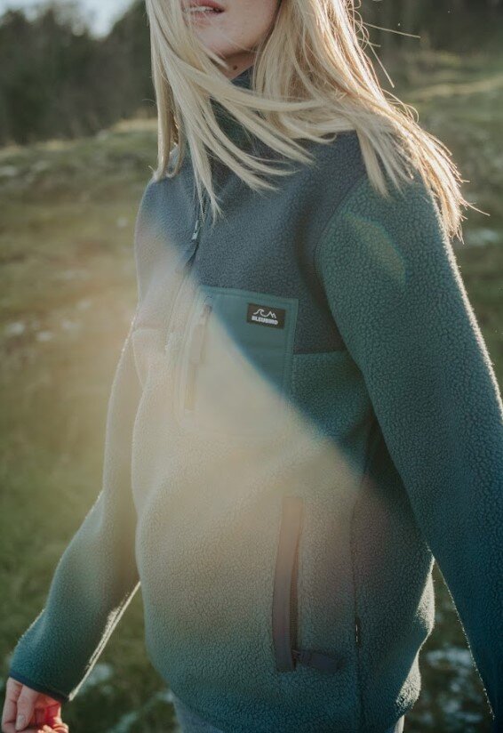 A woman wearing a dark teal fleece pullover jacket with a small black Bleubird logo patch on the chest, photographed outdoors in a grassy field with warm sunlight and lens flare.