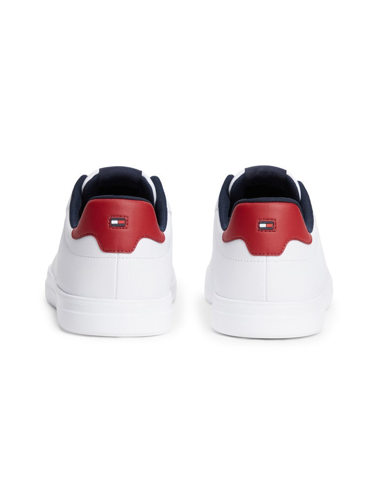 A pair of white low-top sneakers shown from the back, featuring a red heel tab with a small Tommy Hilfiger flag logo and navy blue laces and collar lining.