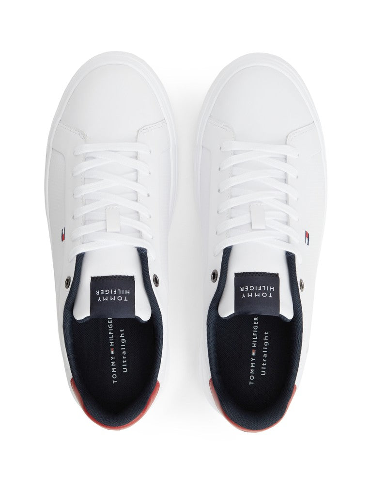 A pair of white low-top sneakers shown from a top-down view on a white background. The shoes feature white laces, a navy tongue label with white text, and a small red and blue flag logo on the side.