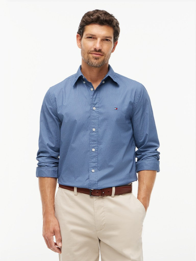 A man wearing a blue Tommy Hilfiger long-sleeve button-down shirt with a subtle micro-pattern, paired with beige trousers and a brown belt, against a plain white studio background.