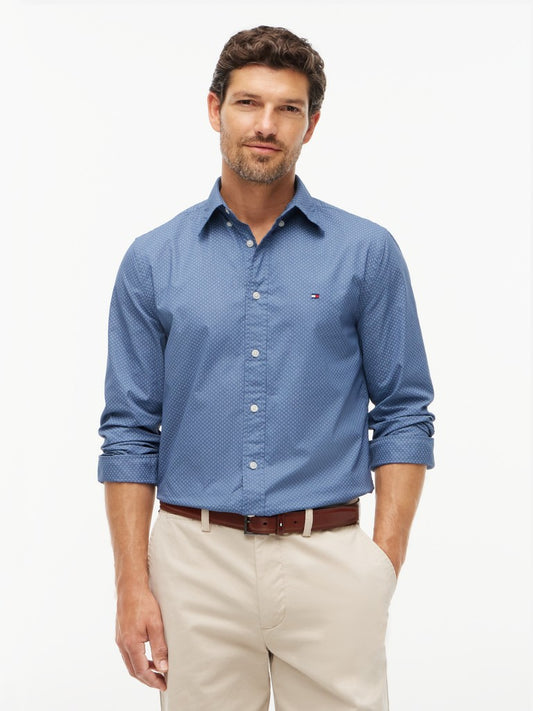 A man wearing a blue Tommy Hilfiger long-sleeve button-down shirt with a subtle micro-pattern, paired with beige trousers and a brown belt, against a plain white studio background.