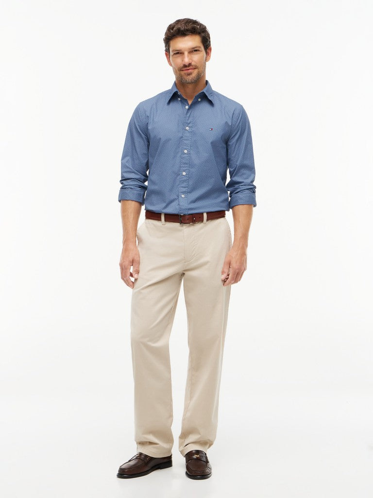 A man modeling a blue long-sleeve button-down shirt with beige trousers, a brown belt, and dark brown leather shoes, photographed against a plain white studio background.