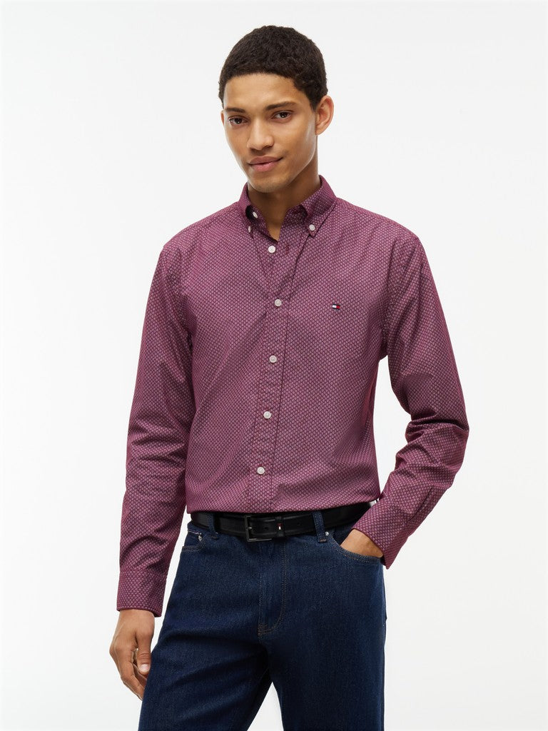 A man wearing a long-sleeve button-down shirt with a small white geometric pattern on a muted purple base, paired with dark blue jeans and a black belt, against a plain white studio background.