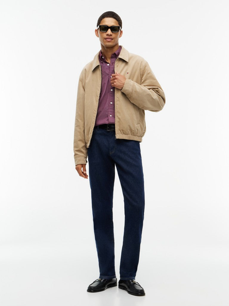 A man modeling a beige zip-up jacket over a purple button-down shirt, paired with navy blue trousers and black leather shoes, against a plain white studio background.
