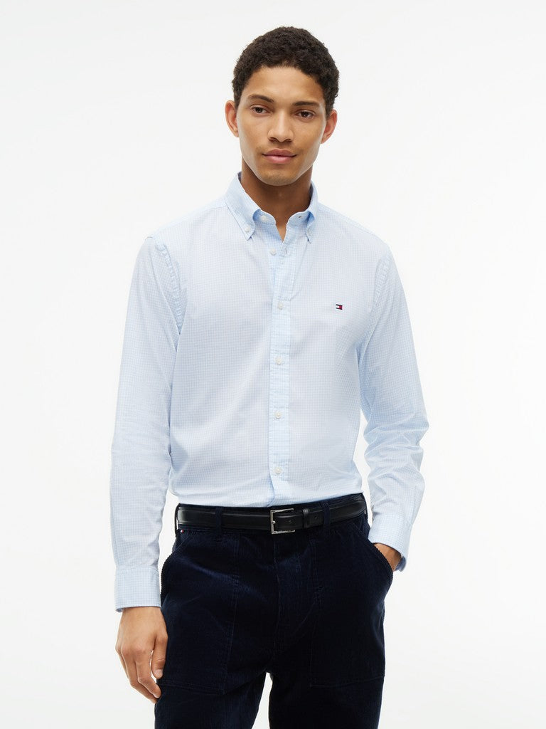 A man wearing a light blue long-sleeve button-down shirt with a small embroidered Tommy Hilfiger flag logo on the chest, paired with dark navy trousers and a black belt, against a plain white background.