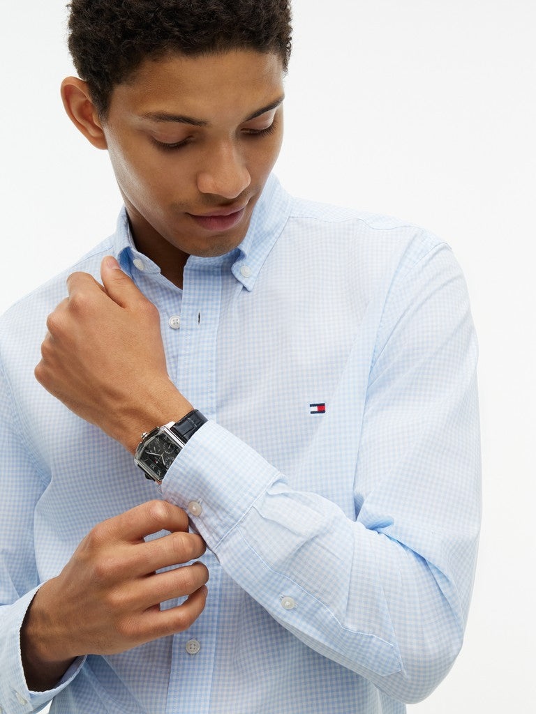 A man wearing a light blue and white checkered button-down shirt by Tommy Hilfiger, adjusting his cuff while looking down. The Tommy Hilfiger flag logo is visible on the chest.