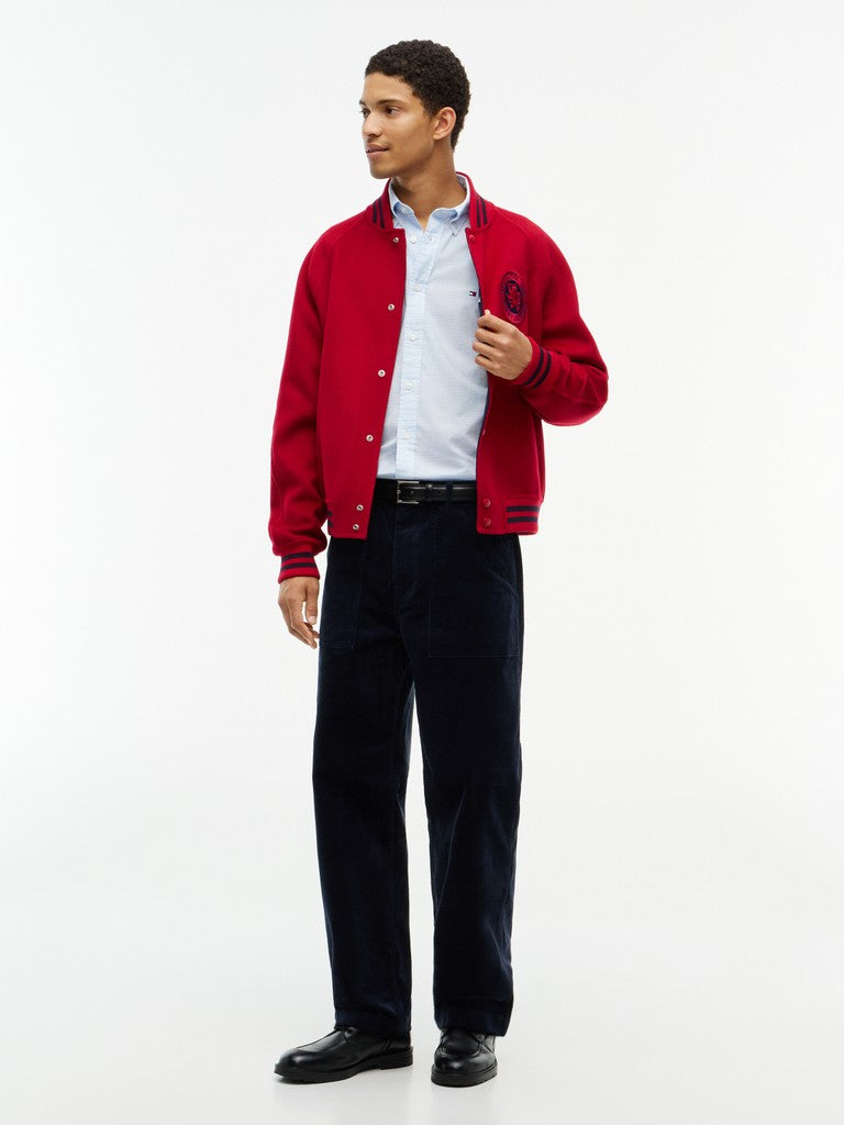 A man modeling a red varsity jacket with navy blue ribbed trim, worn over a light blue dress shirt and dark navy trousers, photographed against a clean white studio background.