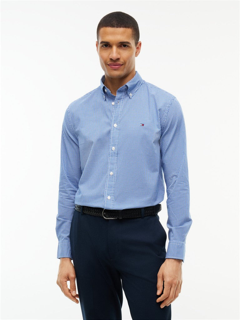A man wearing a light blue and white micro-check patterned button-down shirt by Tommy Hilfiger, paired with dark navy trousers and a black belt, against a plain white studio background.