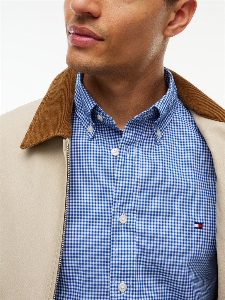 A close-up of a man wearing a blue and white gingham button-down shirt under a beige jacket with a brown corduroy collar and Tommy Hilfiger flag logo