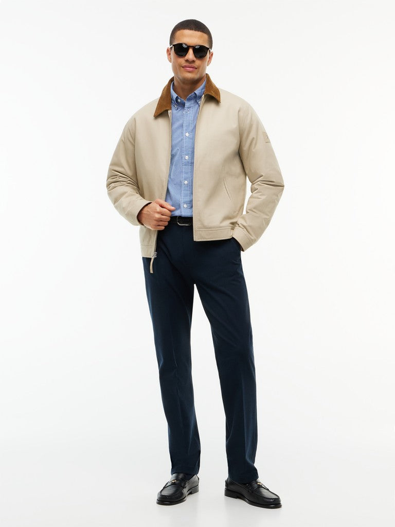 A man modeling a beige bomber jacket with a brown corduroy collar, worn over a light blue button-down shirt and navy trousers, on a white studio background.